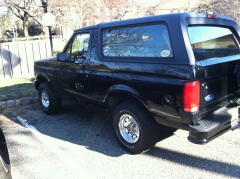 1996 Ford Bronco - A black Ford Bronco is seen from the side, parked in a lot with trees and a fence in the background.