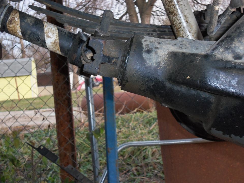Close-up of a vehicle's rear axle with leaf springs and a driveshaft connection. Black and white stripes are visible on a metal tube