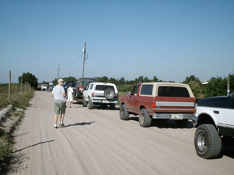 Lifted red and tan Bronco and other trucks parked on a dusty dirt road under blue sky.