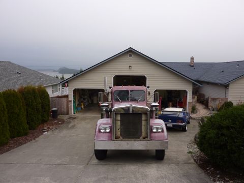 1959 Kenworth W900A - A pink semi-truck is parked in a driveway in front of a garage, with a blue car inside.