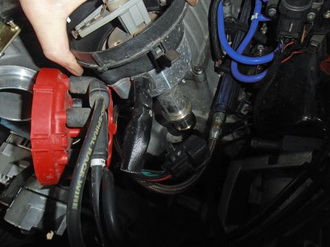 1990 Ford Bronco - Engine bay close-up of a hand tilting a distributor above its mount, exposing the polished shaft and gear end; a red cap fitted with black…