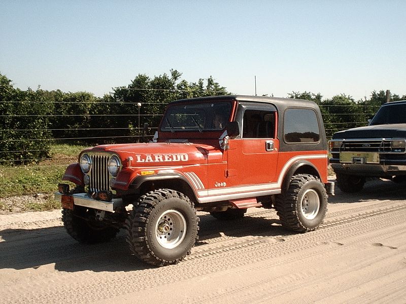 Red Jeep CJ-7 Laredo on mud tires driving on a dirt road past a green hedge