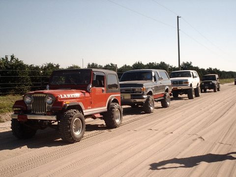 1996 Ford Bronco - A convoy of modified SUVs and Jeeps, including a red Jeep CJ-7 Laredo and several Ford Broncos, drives down a dusty track.