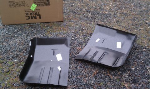 1979 Ford Bronco - Two black car floor pans are shown on a gravel surface, with a cardboard box in the background.