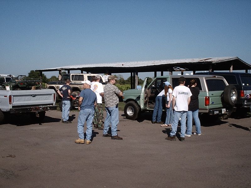 People stand around a green pickup with open door on a dirt lot under an open shelter