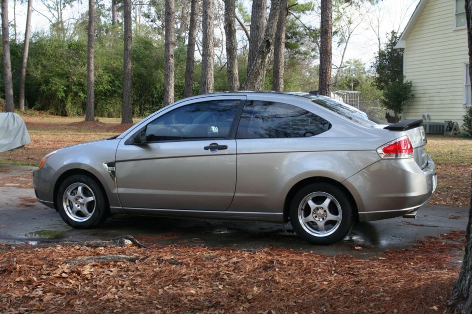 Silver coupe parked on a dirt driveway with trees and a house in the background