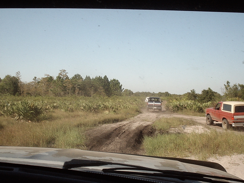 Windshield view of dirt trail with white pickup ahead and red two-tone pickup at right