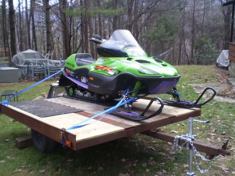 1979 Ford Bronco - A green Arctic Cat snowmobile is strapped to a trailer in a grassy yard with trees in the background.