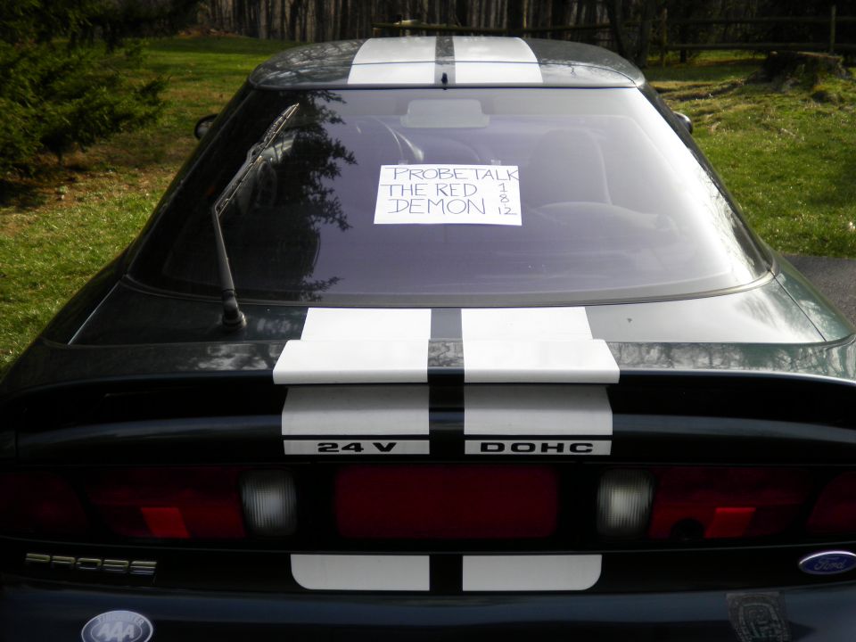 Rear view of a black 1997 Ford Probe with white racing stripes and a note on the back window