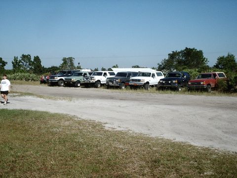 1996 Ford Bronco - Multiple Ford Broncos and trucks are parked in a line on a dirt lot under a clear blue sky.