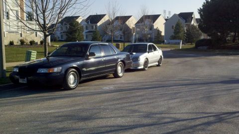 1999 Mercury Grand Marquis - Two cars are parked on a street in front of houses, one is a dark sedan and the other is silver.