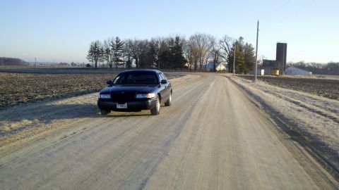 1999 Mercury Grand Marquis - A dark sedan travels down a rural dirt road with fields and farm buildings in the background.