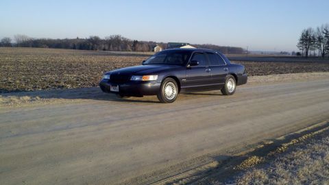 1999 Mercury Grand Marquis - A 1999 Mercury Grand Marquis sits on a dirt road with a field in the background under a clear sky.