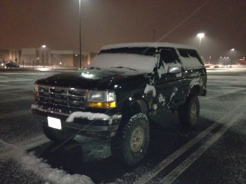 1995 Ford Bronco - Dark green 1995 Ford Bronco covered in snow, parked in a lot with streetlights visible.