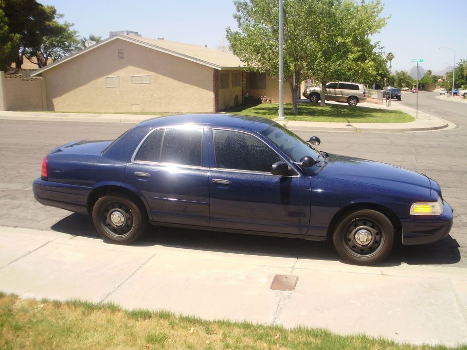 Dark blue Crown Victoria sedan with black steel wheels and spotlight, parked curbside in a suburban neighborhood
