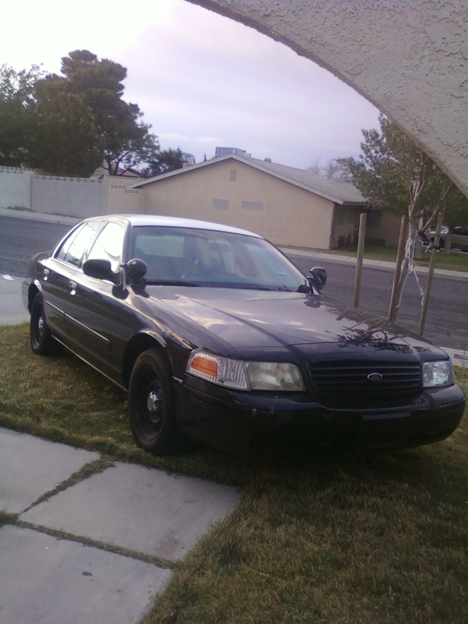 Black Ford Crown Victoria with black steel wheels and A-pillar spotlights parked on grass, front 3/4 view