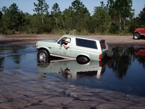 1996 Ford Bronco - A 1996 Ford Bronco is partially submerged in a muddy puddle, with another red vehicle visible in the background.