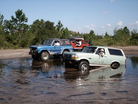1996 Ford Bronco - Three off-road vehicles navigate through a muddy puddle under a clear blue sky.
