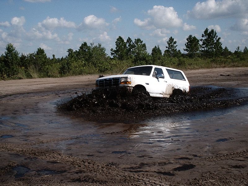White Ford Bronco SUV churning through a muddy off-road pit, spraying mud.