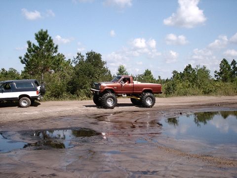 1996 Ford Bronco - Two off-road vehicles are parked on a dirt road next to puddles under a partly cloudy sky.