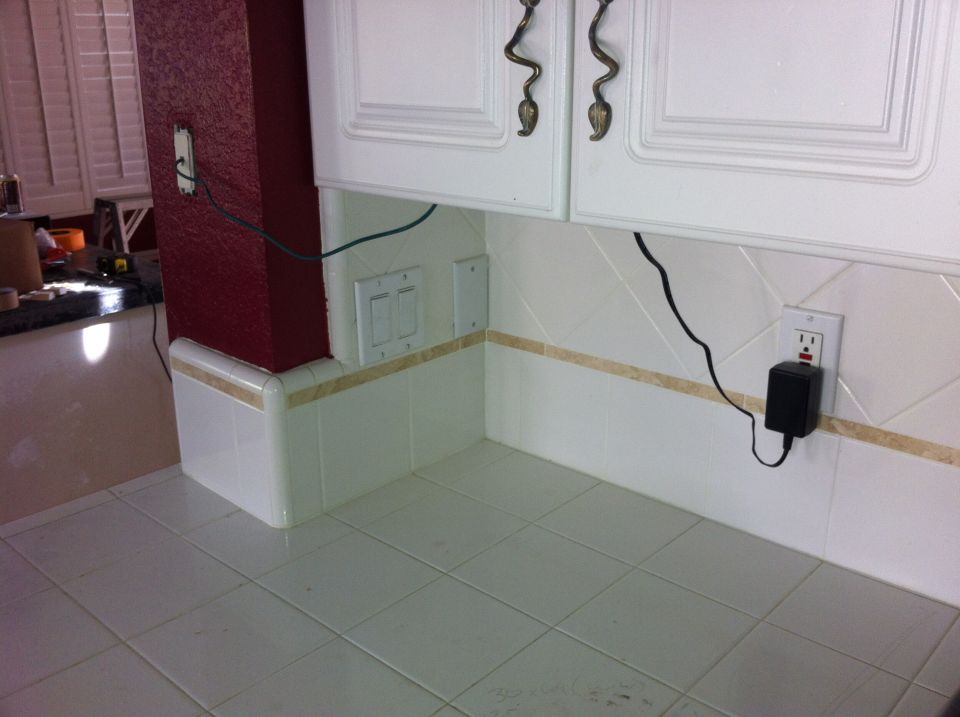 Kitchen counter with white tiled backsplash, red accent wall, and white cabinets. Electrical outlets and a black power adapter are visible