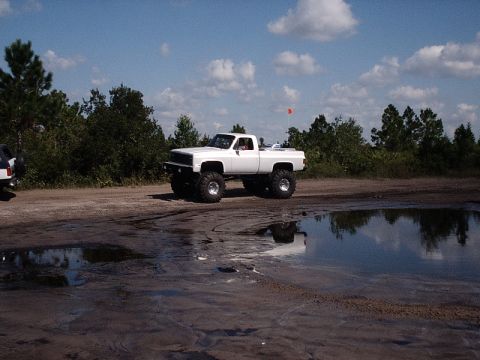 1996 Ford Bronco - A lifted white pickup truck sits on a muddy track under a partly cloudy sky.