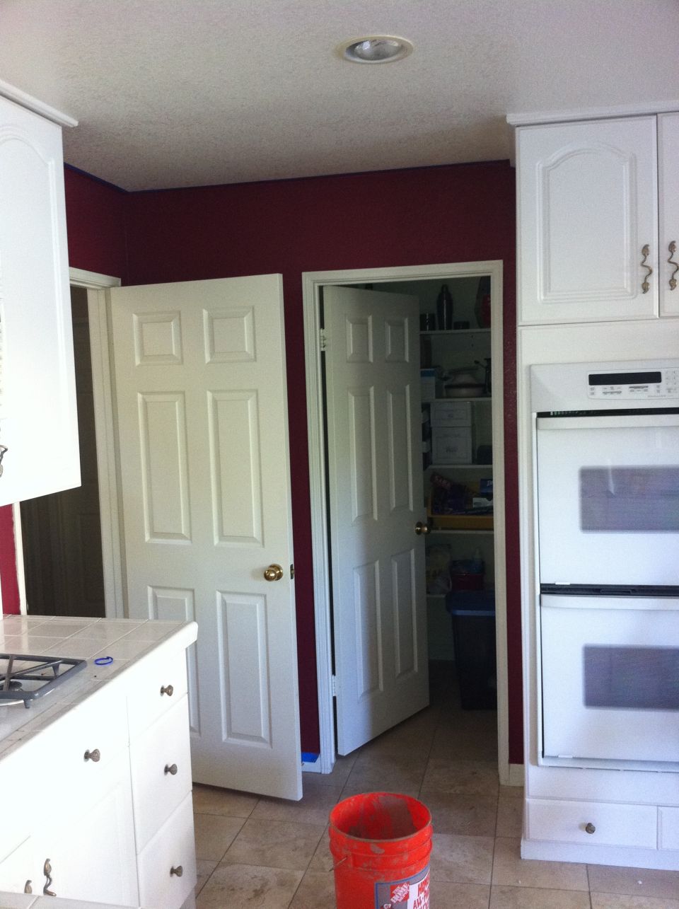Kitchen interior with white cabinets, double ovens, and two doors, one open to a pantry