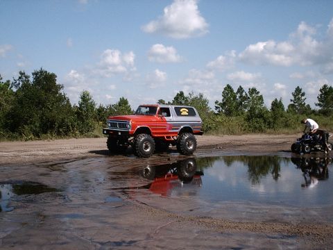 1996 Ford Bronco - A lifted, two-tone SUV sits in a muddy puddle reflecting the sky, with a person on an ATV in the distance.