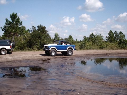 1996 Ford Bronco - Two SUVs, including a 1996 Ford Bronco, are parked on a dirt road near trees and puddles.