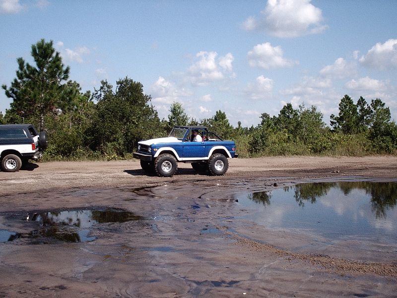 Blue and white lifted early Ford Bronco on sandy off-road trail near puddles