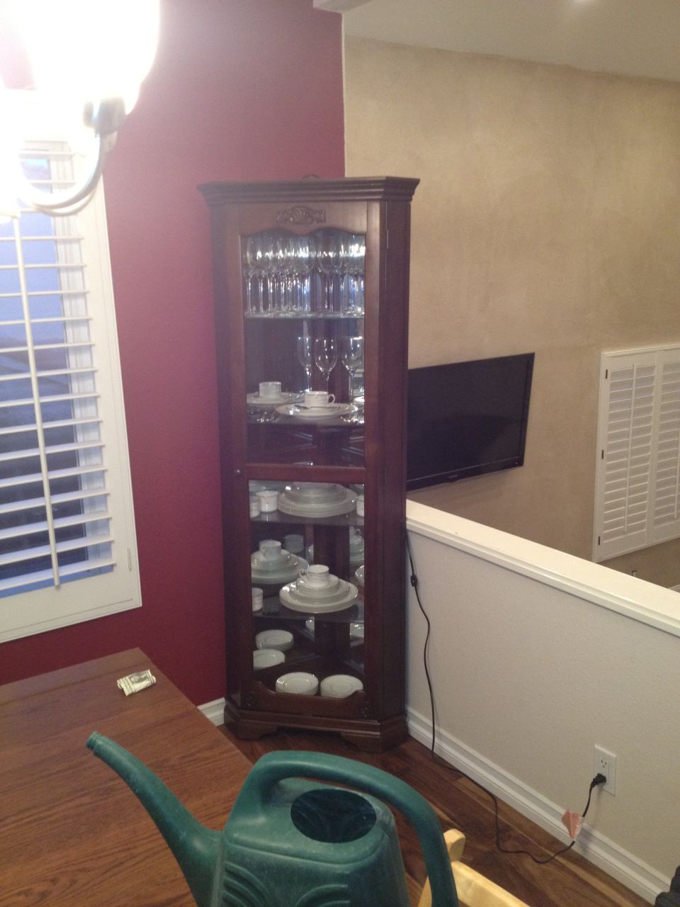 Corner curio cabinet filled with plates, bowls, and glasses. A watering can sits on a wooden table in the foreground