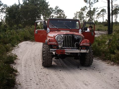 1996 Ford Bronco - A red Jeep CJ is parked on a sandy dirt road surrounded by vegetation and trees.