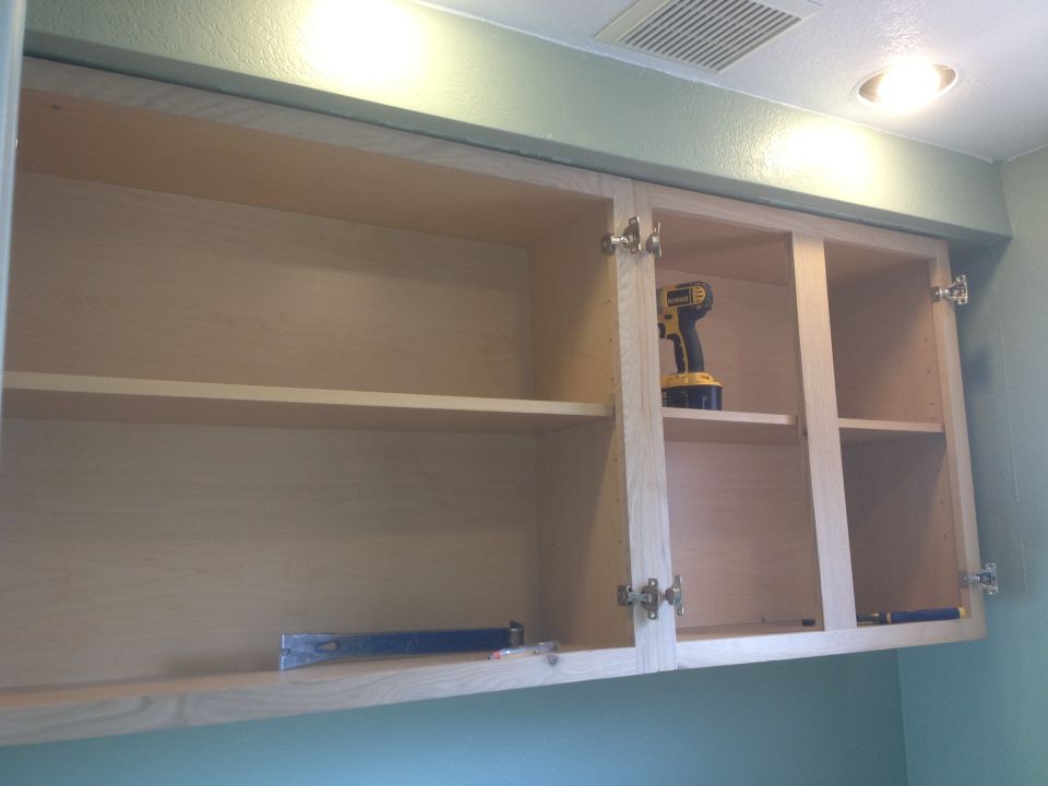 Empty kitchen cabinets with a drill on a shelf and a pry bar on the bottom shelf