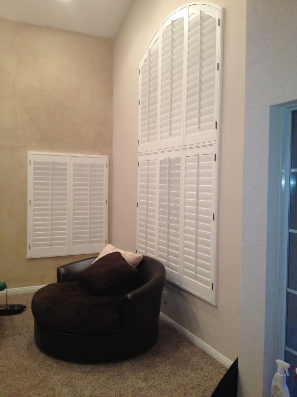 Interior shutters on a large arched window and a smaller rectangular window, with a dark brown armchair and pillows in the foreground