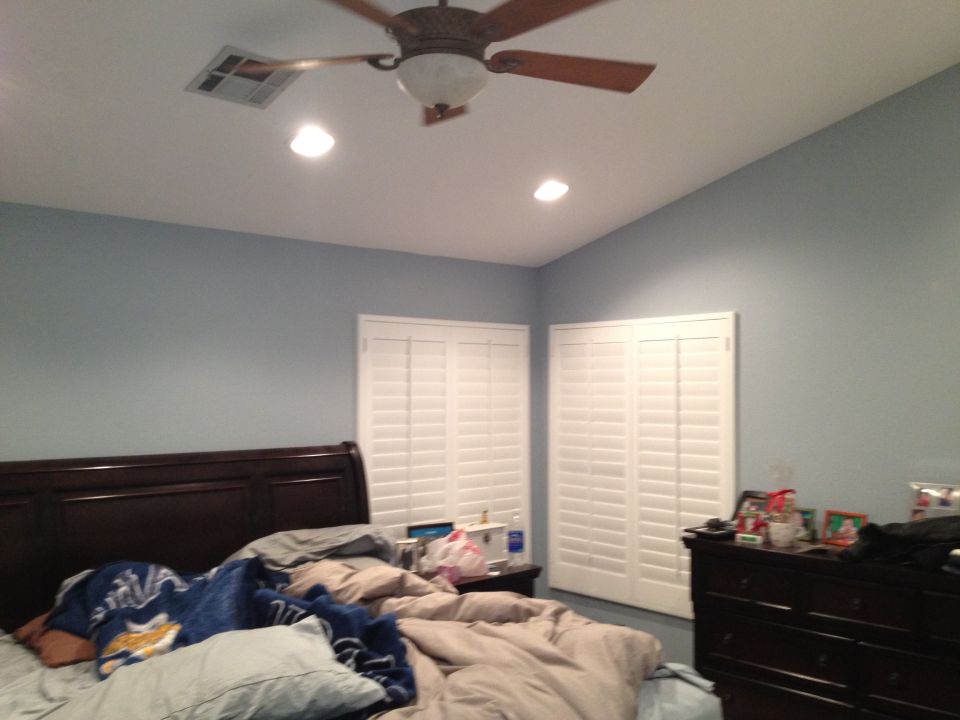 Bedroom with unmade bed, dark wood headboard, blue walls, white plantation shutters, ceiling fan, and dresser
