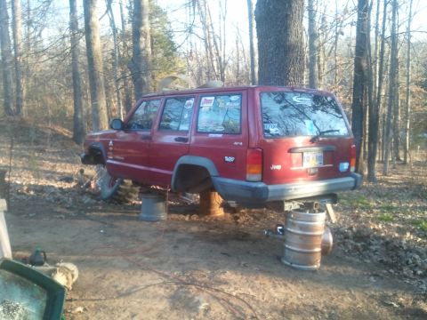 1978 Ford Bronco - Jeep Cherokee XJ being worked on, raised on barrels in a forest setting.