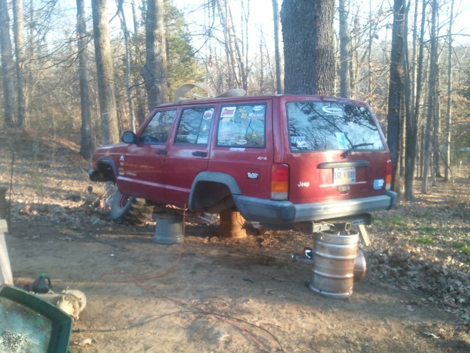 Red Jeep Cherokee XJ, lifted on barrels in a wooded area. License plate visible