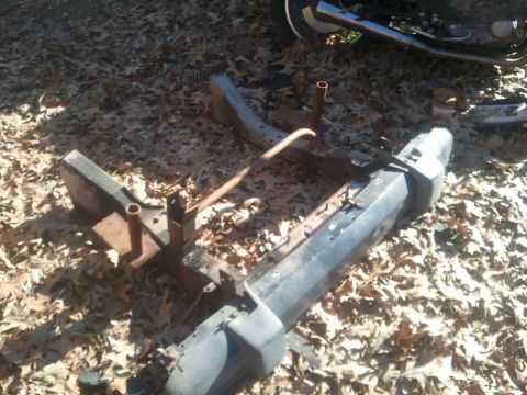 1978 Ford Bronco - Various vehicle parts, including a front bumper and frame components, lie scattered on a bed of fallen leaves.