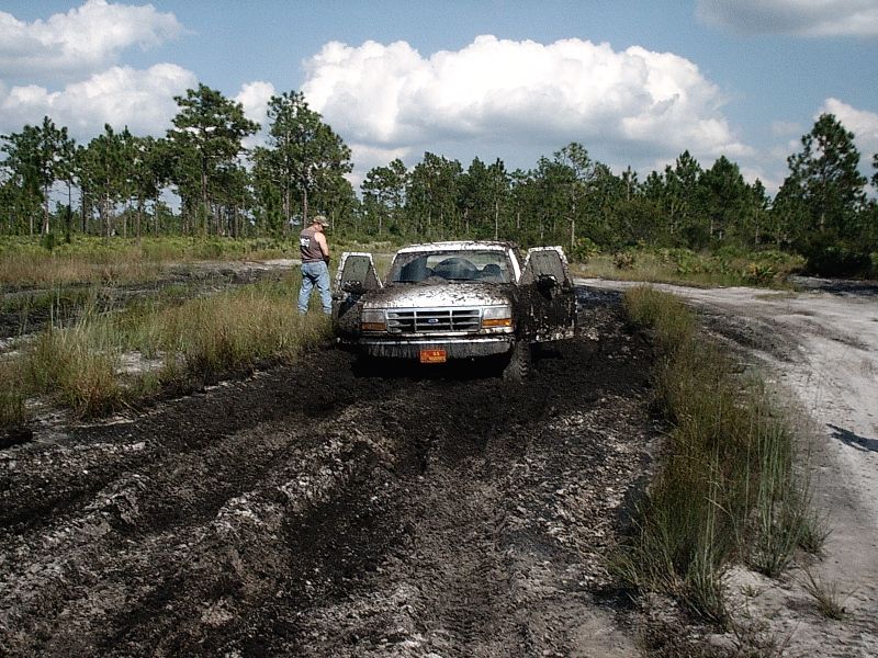 White Ford truck covered in mud, doors open, stuck on rutted off-road trail with a man standing beside it.