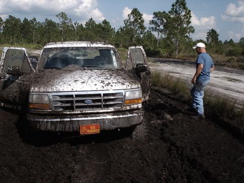 1996 Ford Bronco - A man stands by a white Ford F-Series truck deeply stuck in mud with its doors open.
