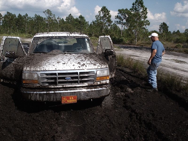 Mud-covered white Ford stuck in deep mud, front view, man standing beside it