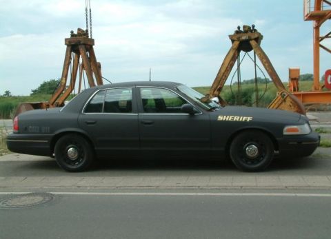 2003 Ford Crown Victoria - A dark-colored Ford Crown Victoria, marked as a sheriff's vehicle, is parked outdoors near large industrial machinery.