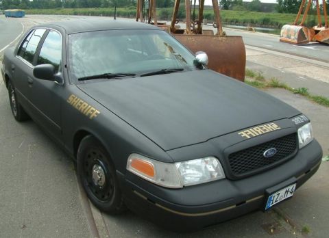 2003 Ford Crown Victoria - A matte black 2003 Ford Crown Victoria police car, marked "SHERIFF", is parked outdoors near industrial equipment.