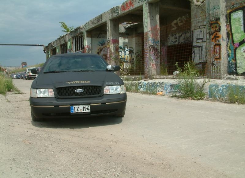 Front view of a black Ford Crown Victoria with 'SHERIFF' on the hood, parked in front of a graffiti-covered building