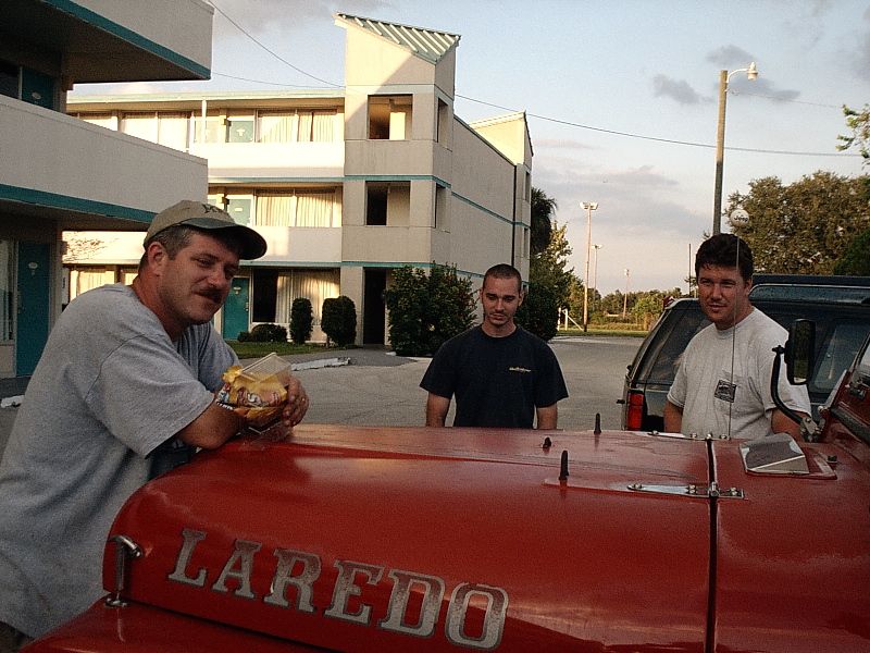 Red vehicle with LAREDO hood lettering and three men at a motel parking lot