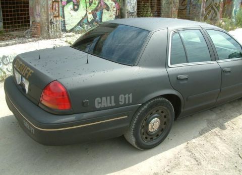 2003 Ford Crown Victoria - Rear quarter view of a matte black 2003 Ford Crown Victoria police car with 'SHERIFF' and 'CALL 911' markings.