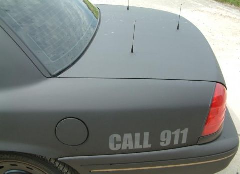 2003 Ford Crown Victoria - This 2003 Ford Crown Victoria, likely a police interceptor, features a matte black finish and multiple antennas on its roof.