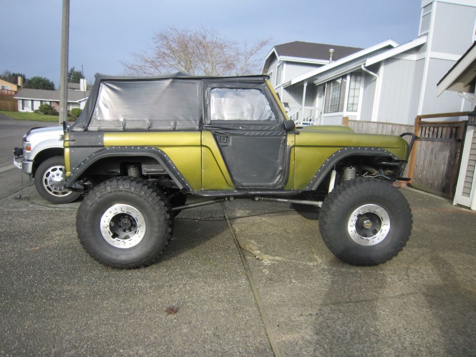1974 Ford Bronco modified with large off-road tires and a soft top, parked next to a pickup truck