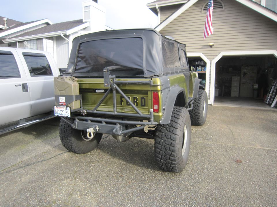 Green Ford Bronco with soft top, spare tire carrier, and gas can mounted on rear. Parked next to a silver pickup truck in a driveway