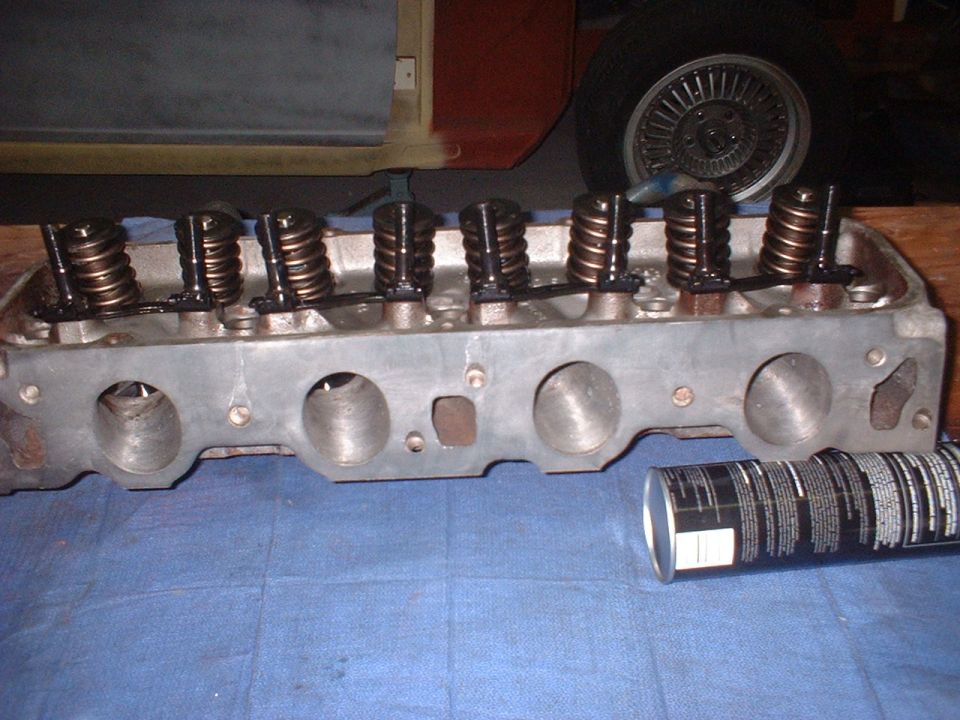 Engine cylinder head with valve springs and retainers, next to a can of lubricant. A car wheel is visible in the background